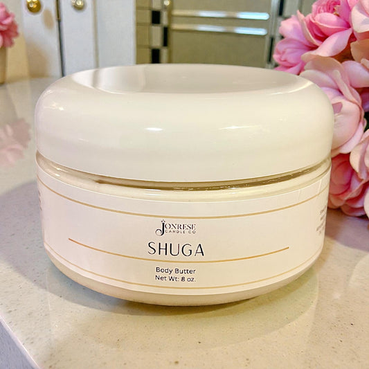 White jar of Jonrese Shuga Body Butter on a marble surface with pink flowers in the background