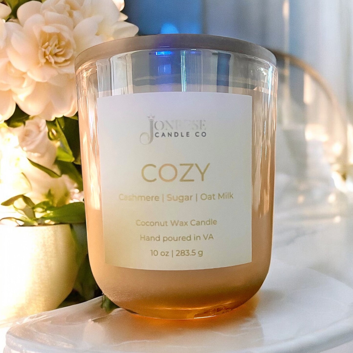 Champagne colored candle jar with 'Cozy' label by Joneise Candle Co on a surface with flowers in the background