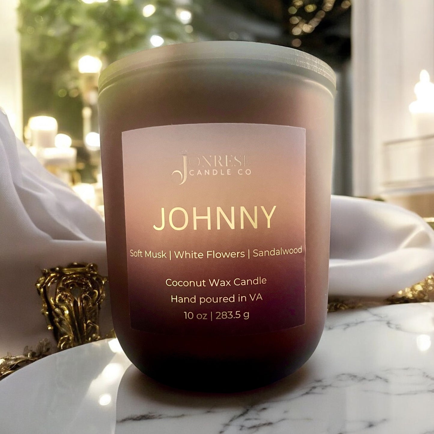 Matte Amber Candle labeled 'JOHNNY' on a marble surface white silk fabric, lights and greenery in the background.