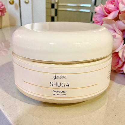White jar of Jonrese Shuga Body Butter on a marble surface with pink flowers in the background