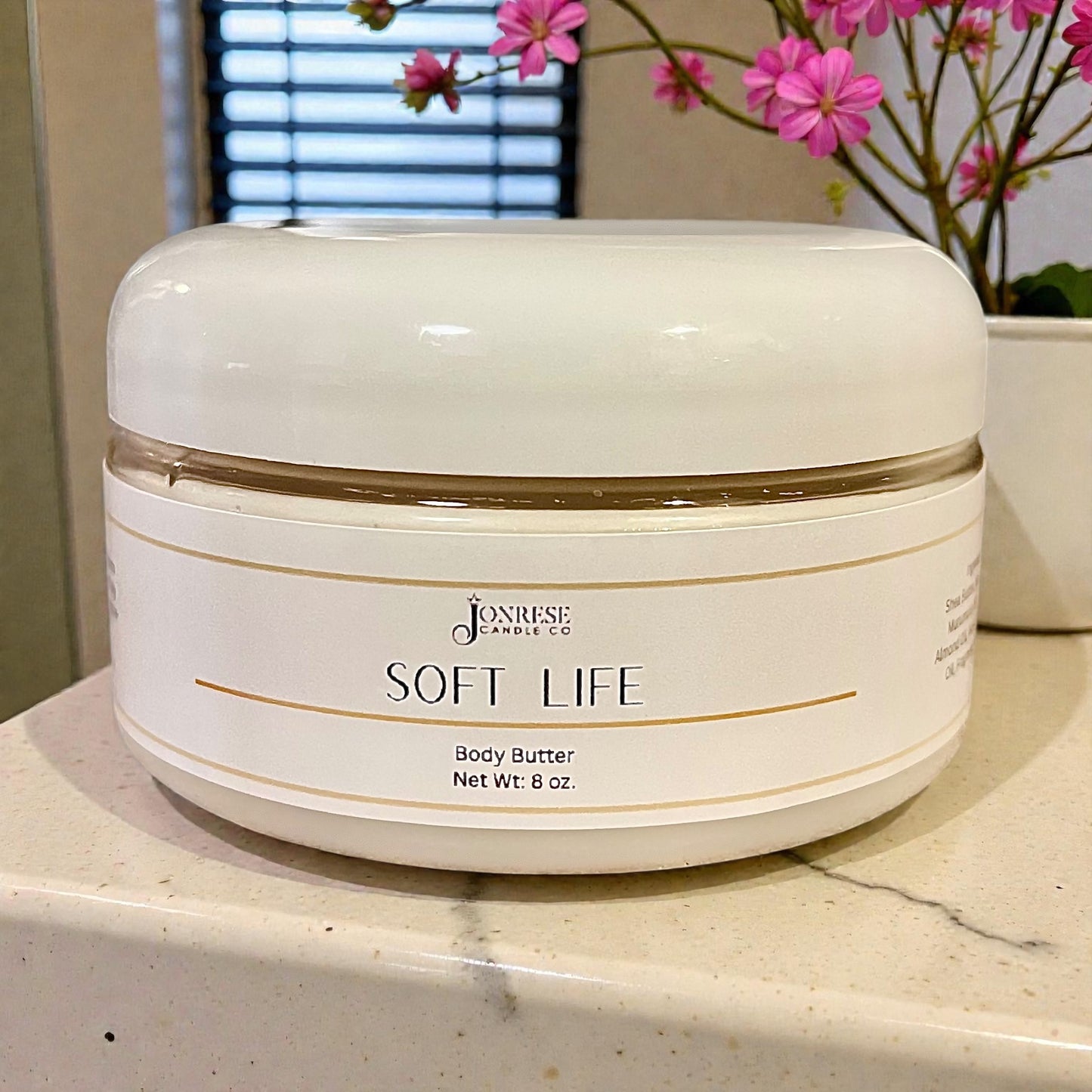White jar of 'Soft Life' body butter on a countertop with pink flowers in the background