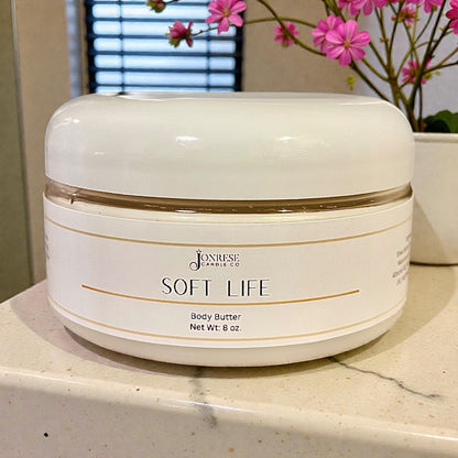 White jar of 'Soft Life' body butter on a countertop with pink flowers in the background