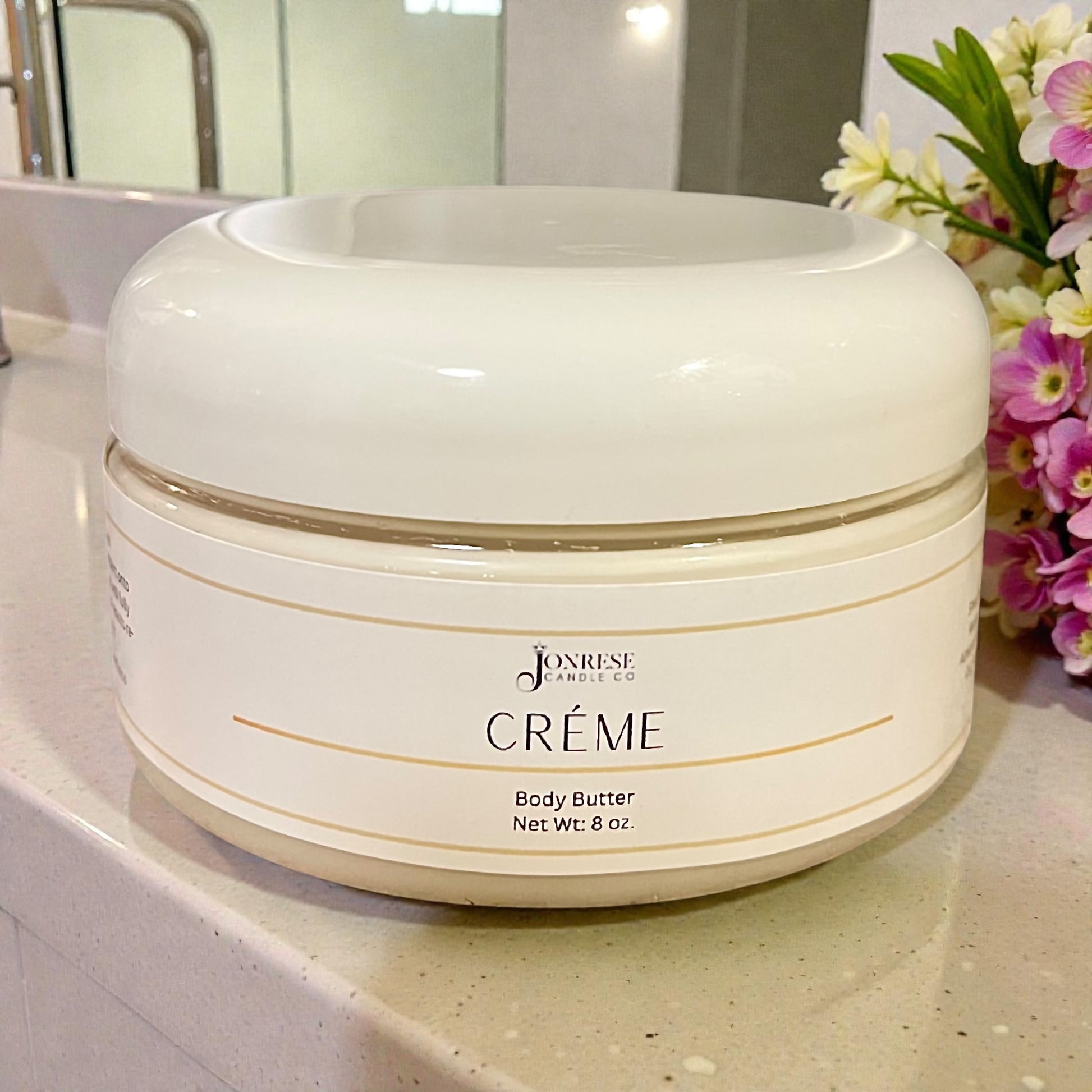 White jar of Jonrese Crème Body Butter on a bathroom counter with flowers in the background