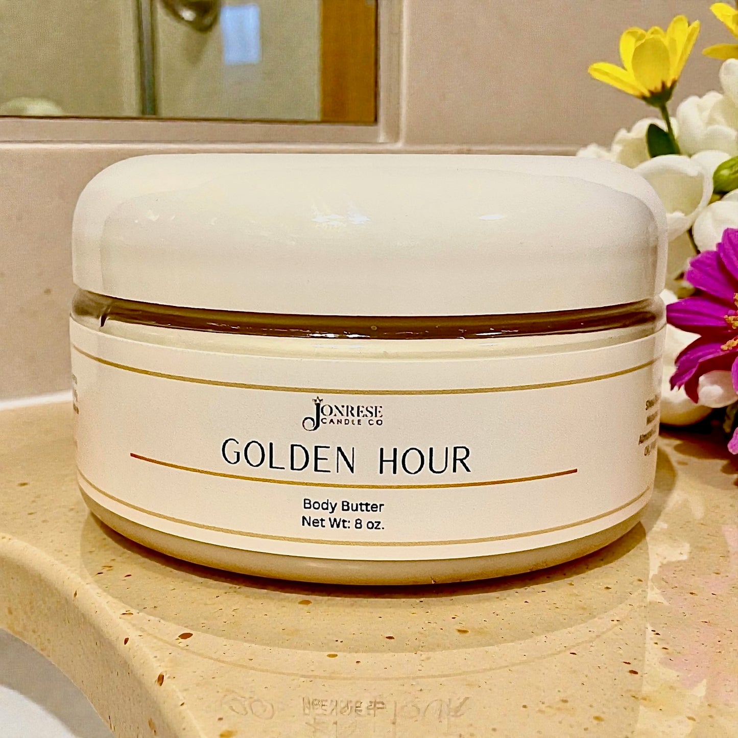 White jar of 'Golden Hour' body butter on a bathroom counter with flowers in the background