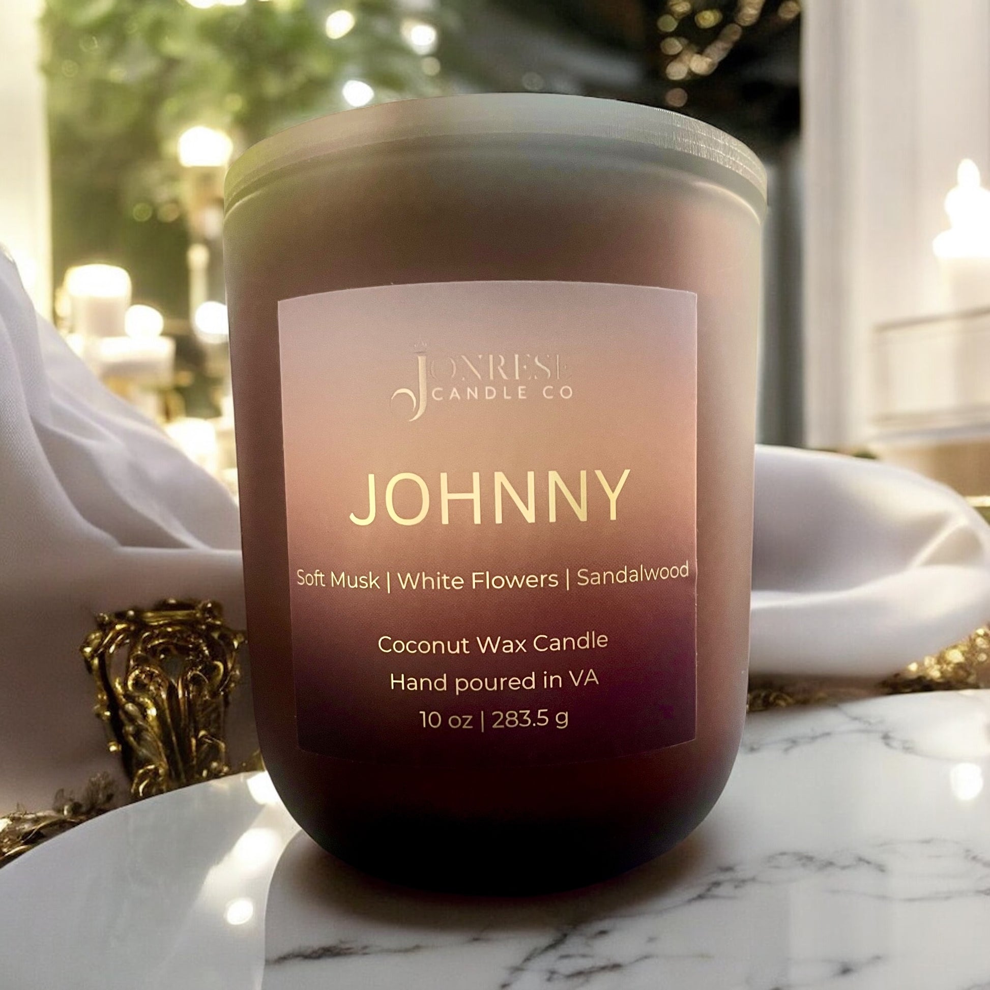 Matte Amber Candle labeled 'JOHNNY' on a marble surface white silk fabric, lights  and greenery in the background.