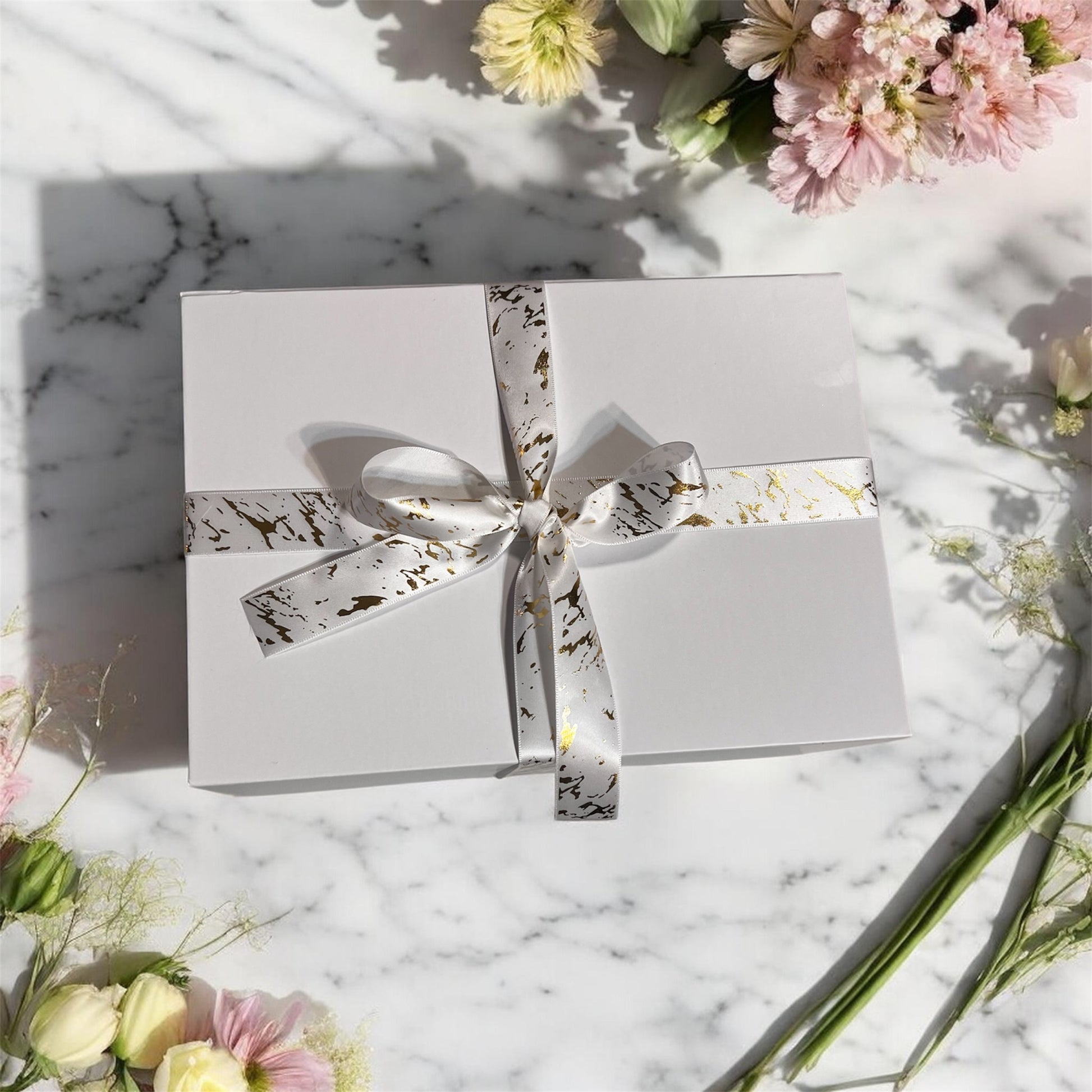 White gift box with a ribbon on a marble surface with flowers around