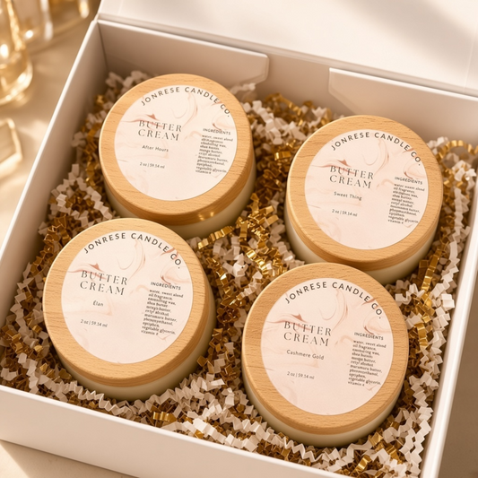 A set of four 2 oz body butter creams in a frosted glass jar with a wood lid and a circle label on the top of the lid sitting in a white gift box on a bed of gold and white crinkle paper