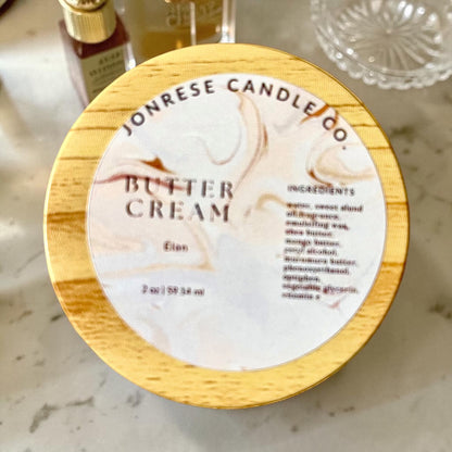 A round jar with a wooden lid for Jonrese Candle Co. Butter Cream Elan scent, featuring a white marble label with elegant brown swirls and black text, set on a light marble countertop.
