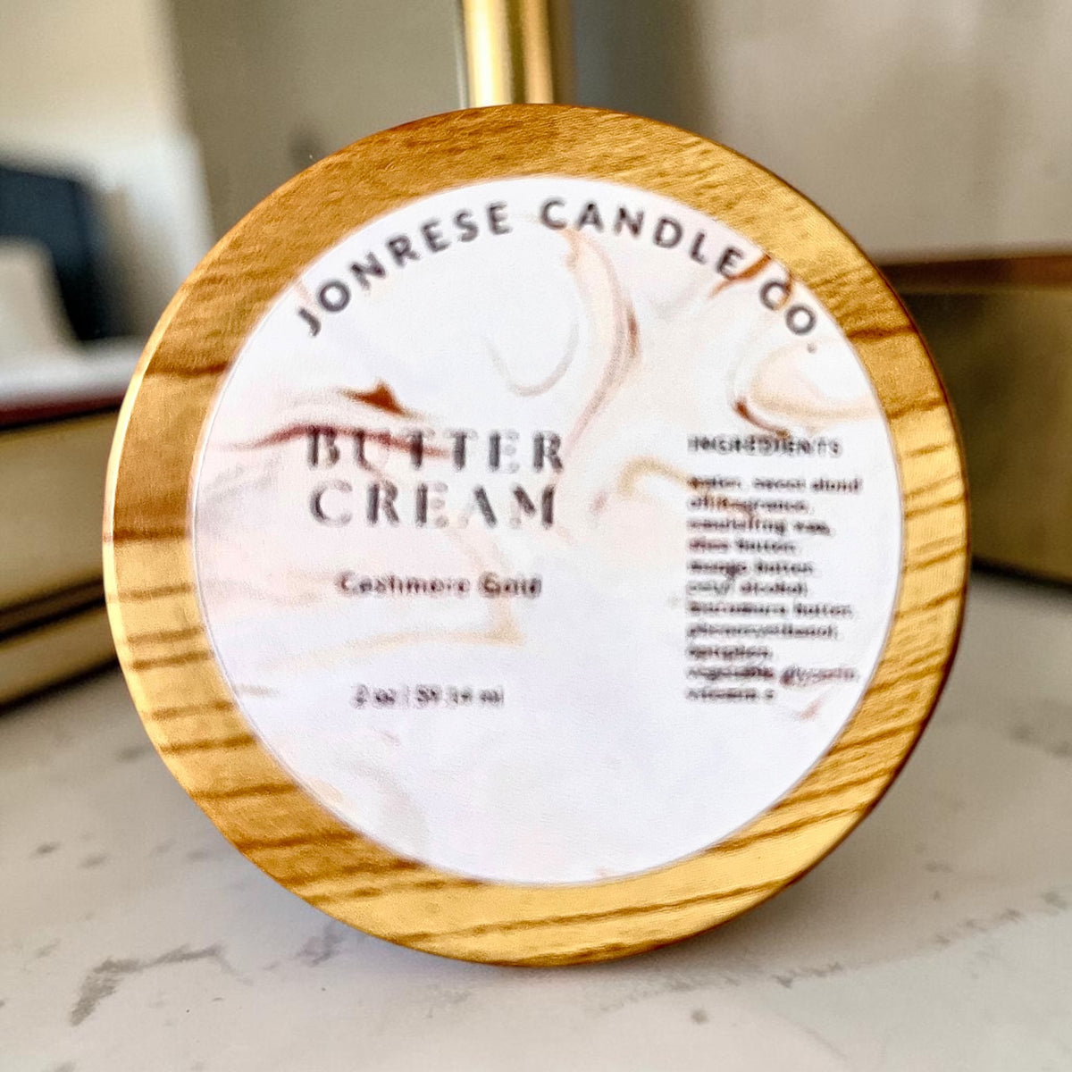 A round jar with a wooden lid for Jonrese Candle Co. Butter Cream Cashmere Gold scent, featuring a white marble label with elegant brown swirls and black text, set on a light marble countertop.