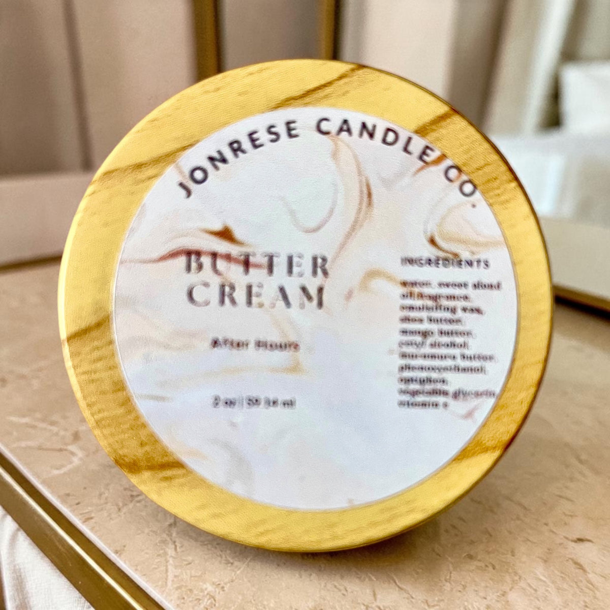 A round jar with a wooden lid for Jonrese Candle Co. Butter Cream After Hours scent, featuring a white marble label with elegant brown swirls and black text, set on a light marble countertop.