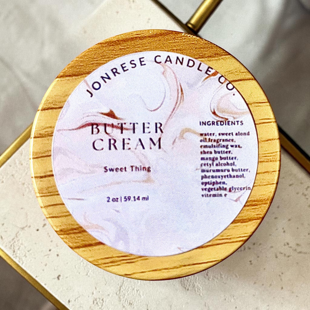 A round jar with a wooden lid for Jonrese Candle Co. Butter Cream Sweet Thing  scent, featuring a white marble label with elegant brown swirls and black text, set on a light marble countertop.