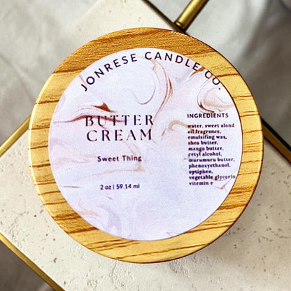 A round jar with a wooden lid for Jonrese Candle Co. Butter Cream Sweet Thing  scent, featuring a white marble label with elegant brown swirls and black text, set on a light marble countertop.