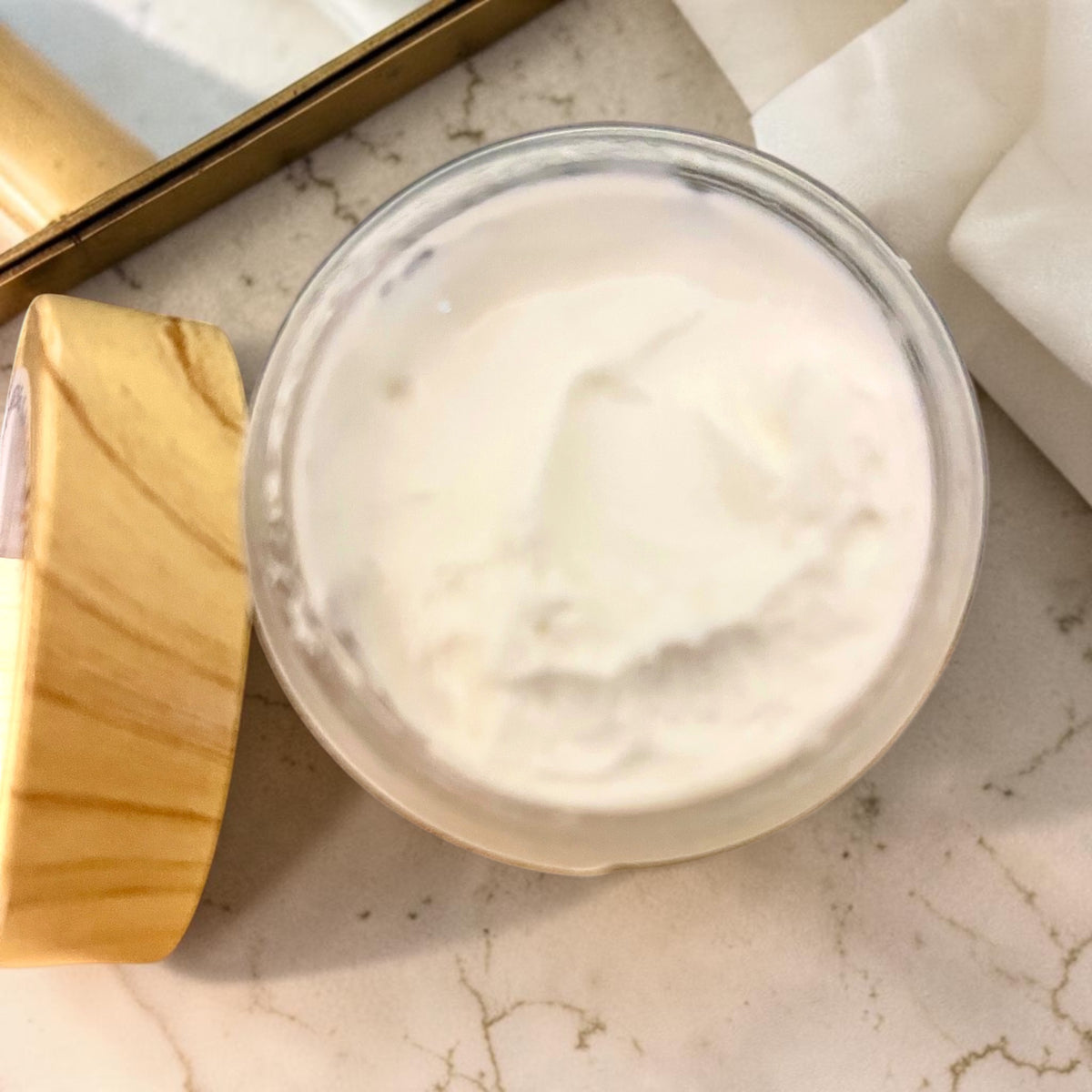 A round open jar with a wooden lid on the side for Jonrese Candle Co. filled with white creamy Butter Cream set on a light marble countertop.