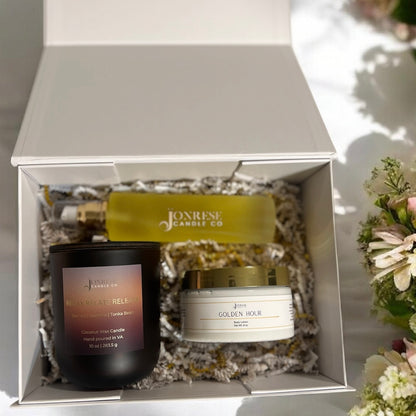 Jonrese Candle Co products including a candle, jar of body lotion and a bottle of body oil in a white gift box with flowers around it.